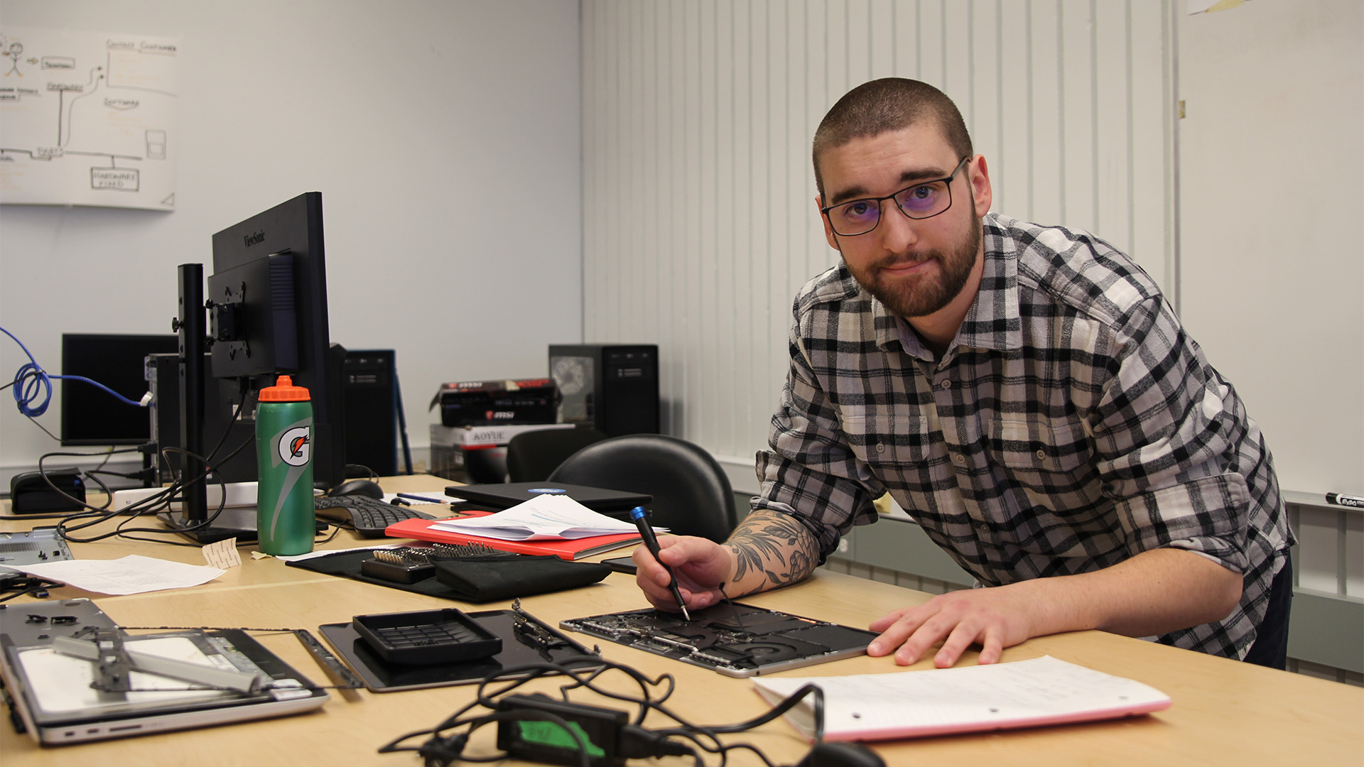 A man in short dark hair, glasses, tattoos and a plaid shirt holding a screwdriver over electronic components of a laptop screen.