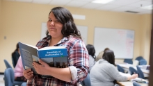 A student standing in a classroom with an open textbook.