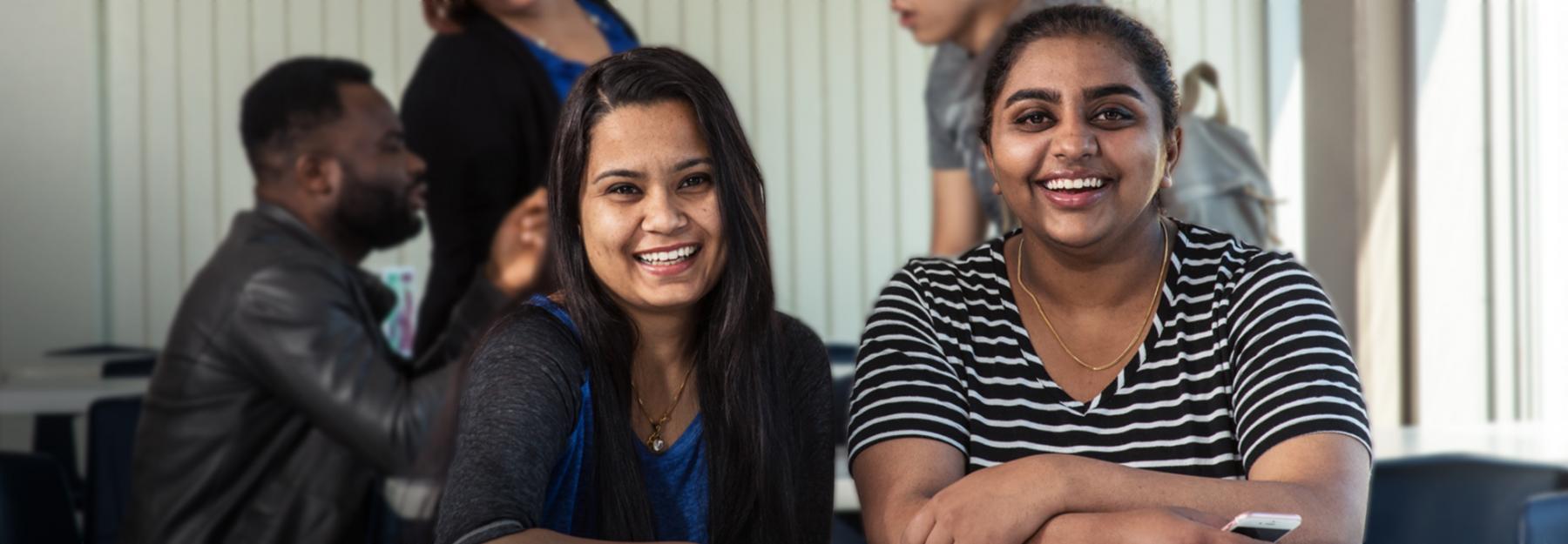Two female international students looking into camera and smiling while in the college cafeteria