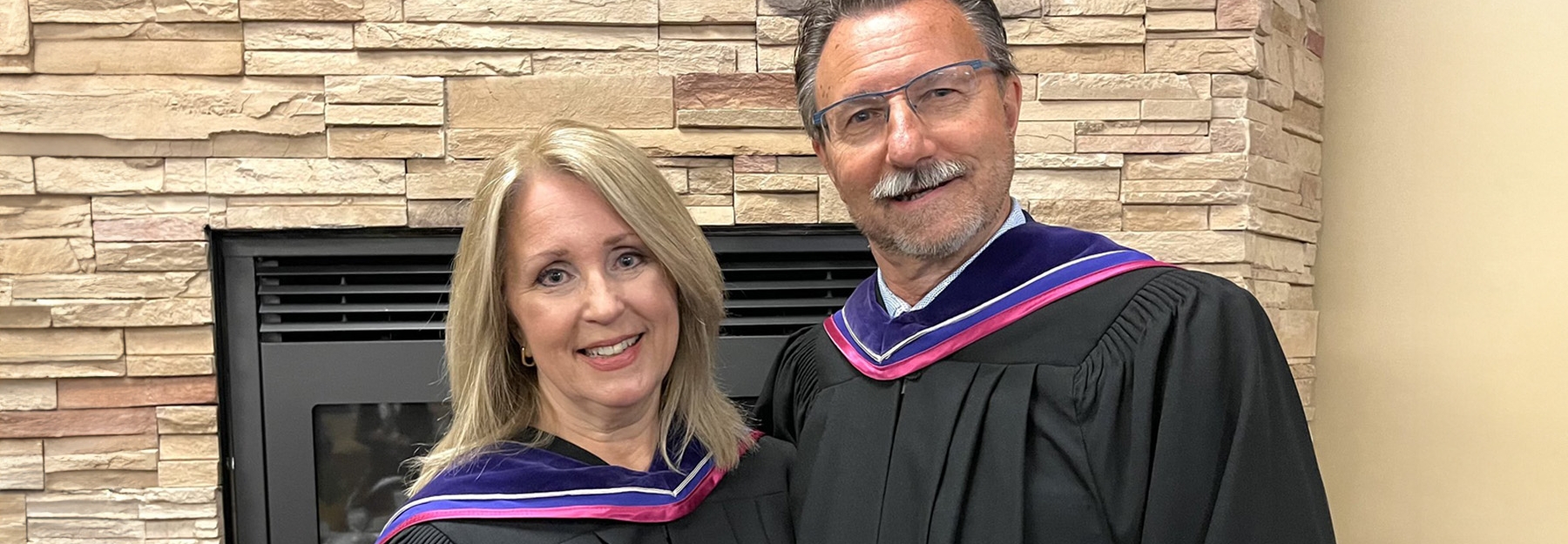 Rick and Karen Chrest standing in black graduation gowns in front of a fireplace.