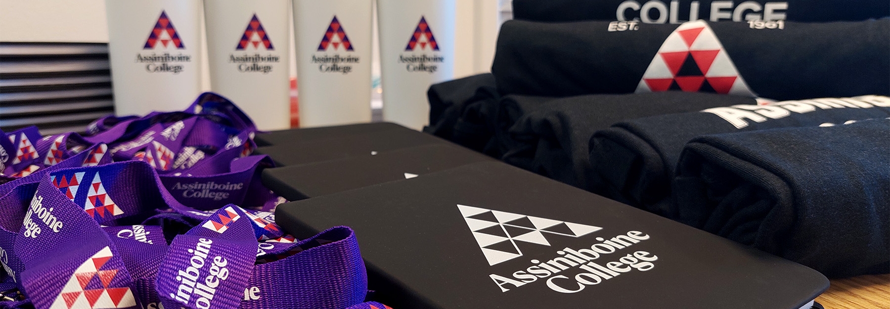 An assortment of branded items with Assiniboine College logos, including purple lanyards, notebooks, sweatshirts and cups.