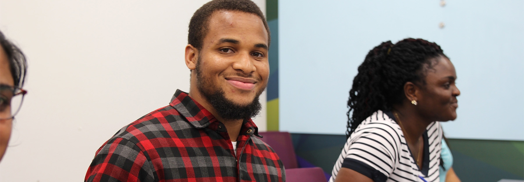 A male student in red plaid shirt smiling at the camera with other students sitting next to him in a classroom.