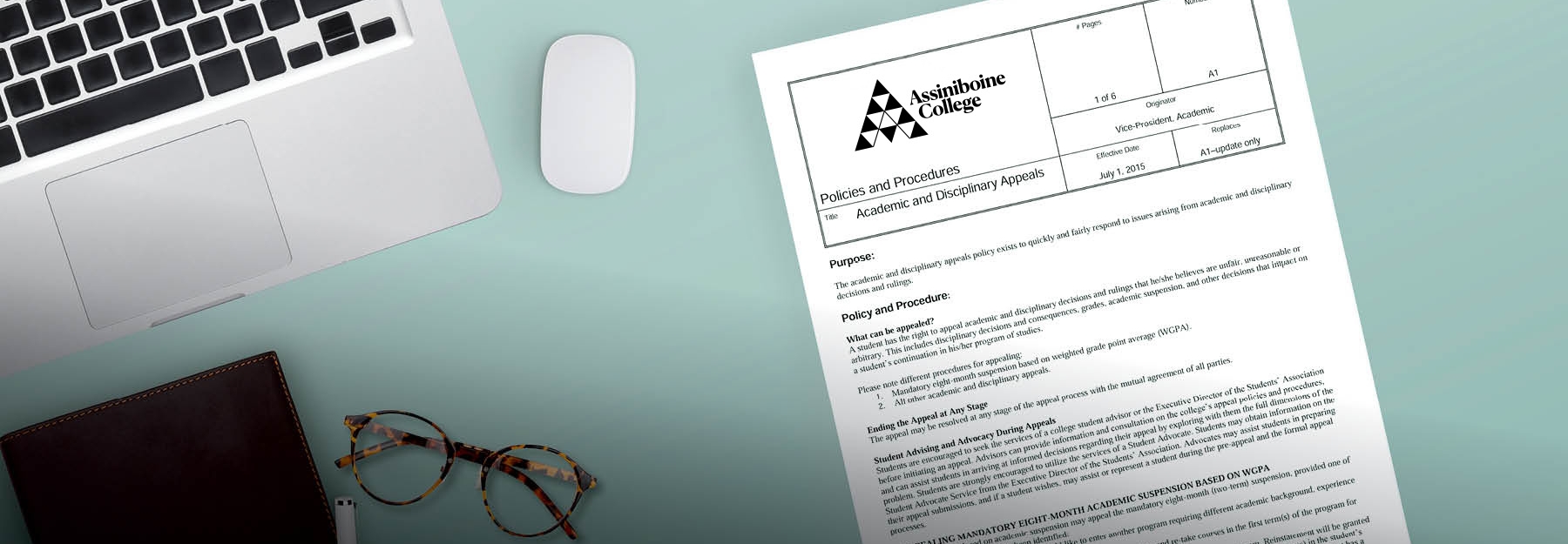 An Assiniboine College policy lying on a teal desk next to a laptop, a pair of glasses and other items.