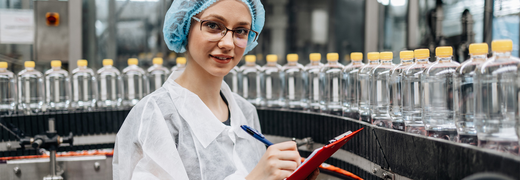 A woman in a white coat and blue hairnet standing with a clipboard in front of a conveyor belt with clear plastic bottles.
