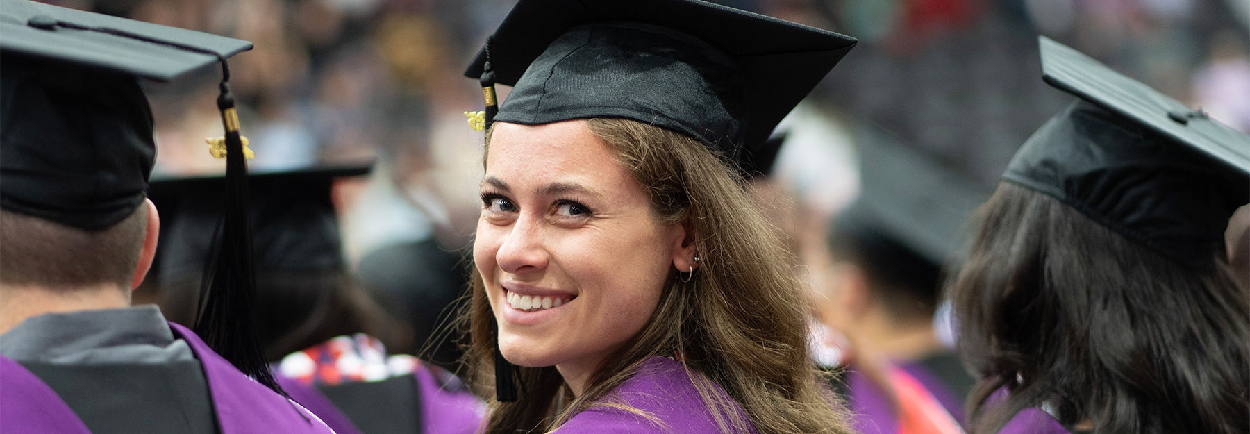 A female graduate in a black gown with a purple-and-red hood looking at the camera while sitting down.