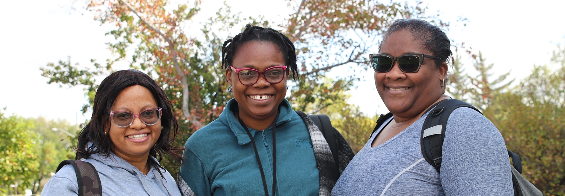 Three women of colour standing together with trees behind them.