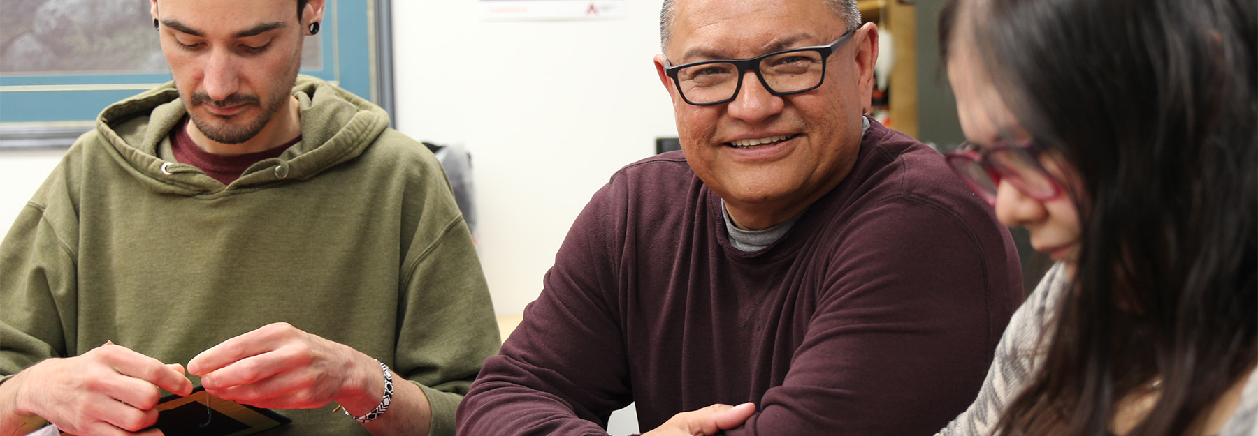 Cecil Roulette sitting at a table in the Cultural Centre with two students working on their beading next to him.