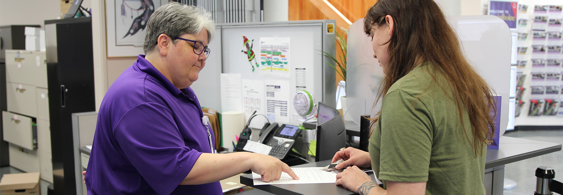 A front-desk staff member in a purple shirt is pointing to a document in front of a prospective student asking for assistance.